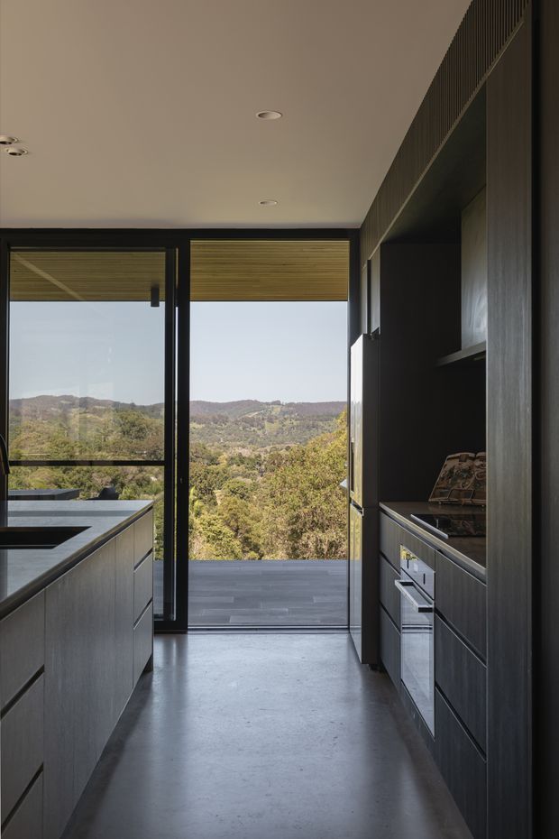 A narrow, contemporary galley kitchen with dark timber-toned cabinetry on both sides, integrated appliances, a central island, and a smooth concrete-look floor. Full-height black-framed glazing opens to a deck or balcony and provides expansive views over a wooded, hilly landscape. The restrained palette and recessed ceiling lights create a sleek, dramatic interior.