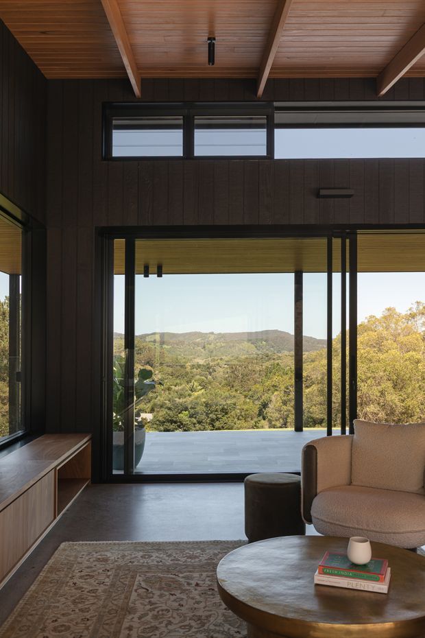 A refined living room with dark vertical timber wall panelling, exposed timber beams and a timber-lined ceiling. Full-height black-framed glazing opens to a broad outdoor terrace and a green, hilly landscape. The room contains a curved upholstered armchair, round coffee table, patterned rug and low built-in timber joinery, creating a warm, contemporary retreat with strong indoor-outdoor connection.