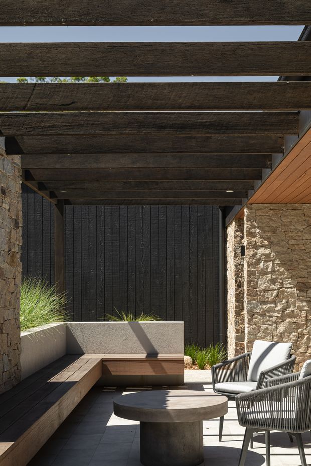 A refined outdoor covered courtyard lounge with a dark timber pergola, exposed beams, rough natural stone walls, black vertical timber screening, and warm timber soffit cladding. Built-in concrete seating and timber benching surround a round concrete coffee table, with woven outdoor armchairs and ornamental grasses adding texture. Strong sunlight creates pronounced shadows across the restrained, earthy material palette.