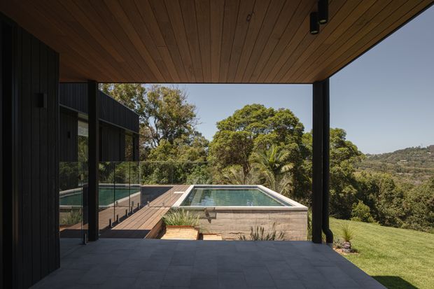 A covered outdoor terrace or patio framed by dark slender columns, with a warm timber-lined ceiling and dark stone paving. Beyond it is a rectangular swimming pool with a raised stone-clad edge, timber decking, glass balustrade, and landscaped grounds overlooking a wooded, rolling hillside. The architecture has a restrained contemporary form with strong indoor-outdoor connection and expansive open glazing.