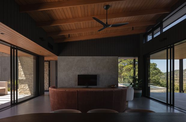 A dramatic contemporary living room with a high timber-lined ceiling and exposed timber beams. A dark sectional sofa faces a simple concrete fireplace or feature wall with a television, while extensive black-framed glazing opens the room to a terrace and leafy landscape. Stone masonry is visible through the glazing, and a ceiling fan hangs centrally.