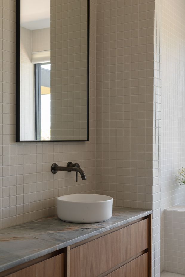 A minimalist bathroom vanity with a round white vessel basin, wall-mounted dark metal faucet, large black-framed mirror, wood-grain cabinetry, and a grey-green veined stone benchtop. Small square pale tiles cover the walls, with a window and partial bathtub visible at the side.