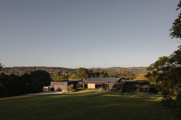 Wide exterior view of a low, single-storey contemporary house set within a broad manicured lawn and surrounded by wooded rolling hills. The building has a horizontally composed form, dark roof planes, large glazing, and muted earthy exterior finishes. Trees frame the foreground and right edge, while the expansive landscape provides a strong sense of seclusion and connection to nature.