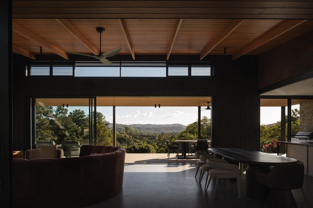 A sophisticated open-plan living and dining area with expansive floor-to-ceiling glazing opening toward a leafy mountainous landscape. The room features a dark, moody interior palette, a warm timber-lined ceiling with exposed structural beams, high-level clerestory windows, dark walls, a large dining table, upholstered dining chairs, and a lounge seating area. A kitchen or bar zone is visible at the right, while the broad opening creates a strong indoor-outdoor connection.