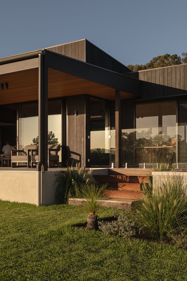 Exterior view of a contemporary single-storey house with dark vertical timber cladding, broad projecting roof forms, black-framed glazing, and a covered outdoor terrace. Timber steps lead from the raised deck to a landscaped lawn with ornamental grasses. Warm late-afternoon sunlight highlights the timber soffit, facade, and outdoor living area.