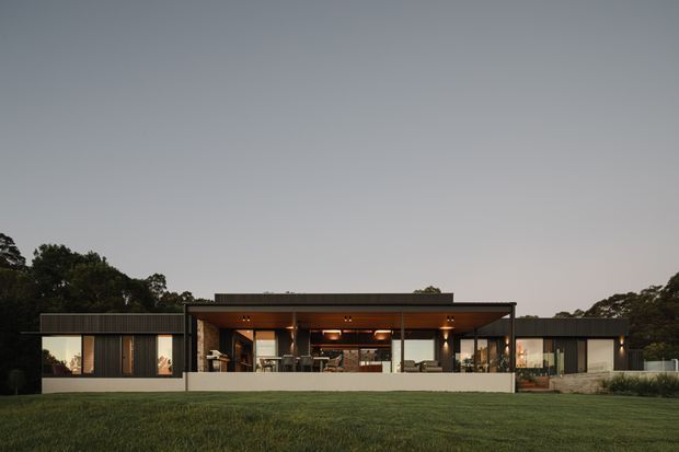A wide, dusk exterior view of a low-slung contemporary single-storey residence set behind an expansive lawn. The house has a long horizontal composition, flat rooflines with broad overhangs, dark vertical cladding, extensive floor-to-ceiling glazing, and a central open living zone with warm timber soffits and illuminated interiors. Trees frame the property in the background.