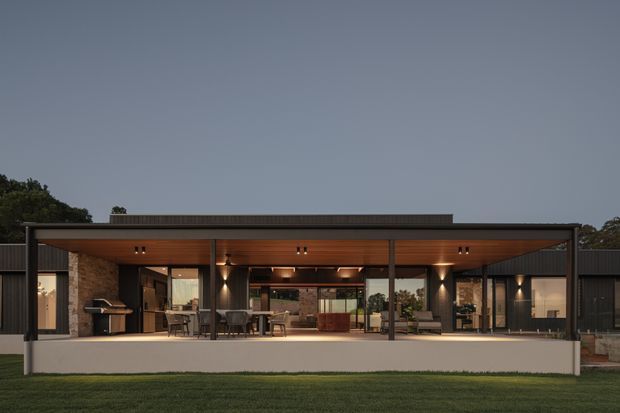 A dusk exterior view of a contemporary single-storey home with a broad covered outdoor entertaining terrace. The dark metal-framed pavilion has a warm timber-lined ceiling, extensive glazing, stone masonry walls, outdoor dining and lounge furniture, and an integrated barbecue area. The building sits on a level landscaped lawn with mature vegetation behind it.