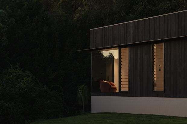A dark, minimalist modern house exterior set against dense greenery. The low-pitched roof and facade are clad in near-black vertical boards, with large dark-framed windows revealing a warmly lit interior and a partial armchair. A pale rendered or concrete plinth forms the base, with a lawn and heavily planted backdrop.