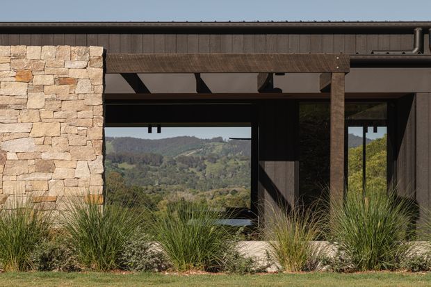 Exterior close-up of a contemporary, dark-clad building with a rustic timber pergola or covered entry framing large windows. A substantial stacked natural-stone wall anchors the facade, while ornamental grasses and low landscaping soften the base. The glazing reflects and frames a wooded, hilly landscape beyond.