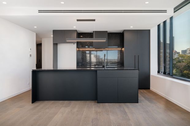 A contemporary open-plan kitchen with a large dark island, integrated dark cabinetry, a reflective splashback, and timber-look flooring. Full-height windows provide city views and abundant natural light, while recessed ceiling lighting and linear air vents contribute to a clean, streamlined fit-out.