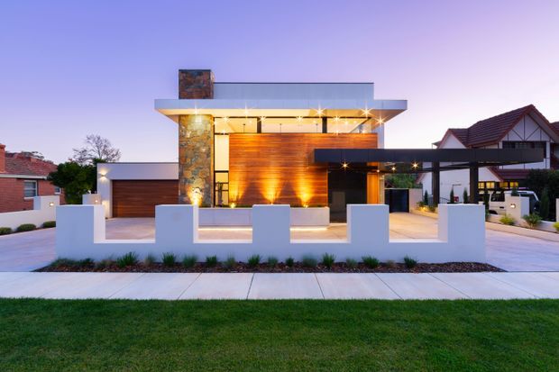 A dusk exterior photograph of a contemporary multi-level residence with a strong horizontal flat-roof form, projecting roof canopy, extensive glazing, warm timber-clad walls, and a prominent natural-stone chimney or feature wall. The front garden includes a low white rendered boundary wall, concrete-paved forecourt, integrated lighting, and restrained planting. Neighbouring suburban homes are visible in the background.