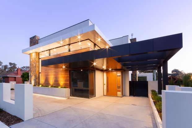 Contemporary multi-level residential facade at dusk, featuring strong horizontal flat-roof forms, expansive glazing, warm timber wall cladding, dark metal fascia and carport canopy, pale rendered walls, and a stone-clad vertical accent. The foreground shows a broad concrete driveway and entry approach, with a covered carport integrated into the building.