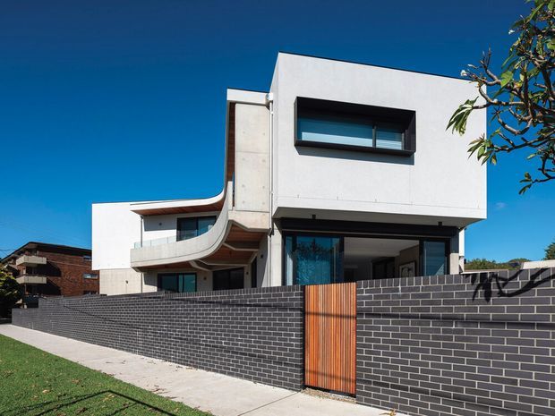A contemporary multi-level residential house with crisp white rendered walls, projecting box forms, dark-framed glazing, and a distinctive curved balcony edge. A long dark brick boundary wall with a warm timber pedestrian gate fronts the property, with neighbouring homes and a clear blue sky in the background.