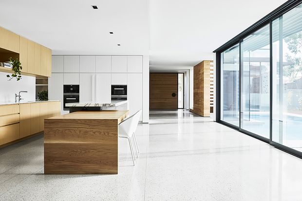 Bright open-plan contemporary interior combining a minimalist kitchen with a timber island and extensive floor-to-ceiling glazing. The kitchen features light wood cabinetry, white built-in cabinetry, integrated appliances, and a white island benchtop. A spacious polished light terrazzo floor extends into the adjoining living or circulation area, with dark-framed glazing overlooking a visible pool or outdoor area.