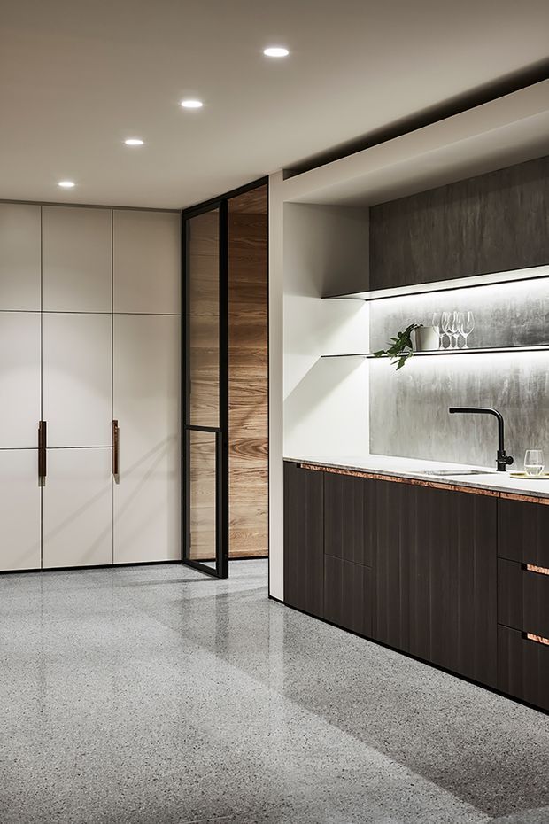 A refined contemporary kitchen with a long dark built-in island and cabinetry, pale integrated pantry cupboards, and open display shelving. The kitchen features a white benchtop, dark textured splashback, black tapware, warm timber detailing, under-shelf lighting, and a polished grey terrazzo-style floor. A black-framed glazed door separates the kitchen from a timber-lined adjoining space.