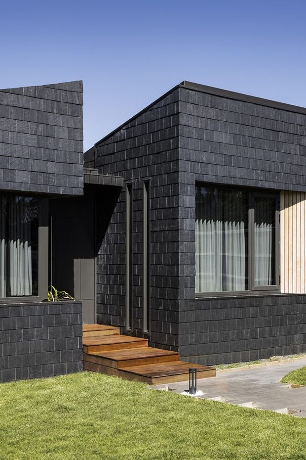 Exterior view of a modern, low-rise residential building composed of angular, box-like volumes. The facade is clad in dark charcoal slate-like rectangular tiles with narrow horizontal coursing, contrasted by warm vertical timber cladding at the right edge and timber entry steps. Dark-framed windows with sheer curtains face a paved walkway and manicured lawn under a clear sky.