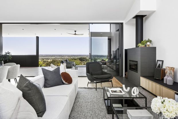 Bright contemporary living room with a white sectional sofa, dark accent armchair, black glass coffee tables, and a large grey rug. A linear black fireplace with timber-toned built-in cabinetry forms the feature wall, while expansive floor-to-ceiling glazing opens to a broad distant landscape view. The room has a clean, refined, open-plan feel with a ceiling fan and strong indoor-outdoor connection.