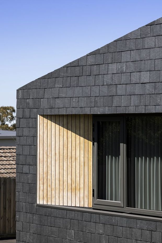 Close-up exterior view of a contemporary building facade clad in dark rectangular slate-like shingles, with a steeply sloping gable roofline. A warm vertical timber screen sits beside a dark-framed glazed window with curtains, creating strong contrast between the black cladding and natural wood.