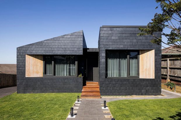 Front exterior view of a compact contemporary house composed of two dark masonry-clad pitched volumes separated by a recessed entry. Large glazed openings have vertical timber screening, with timber steps and a paved path leading across the lawn.