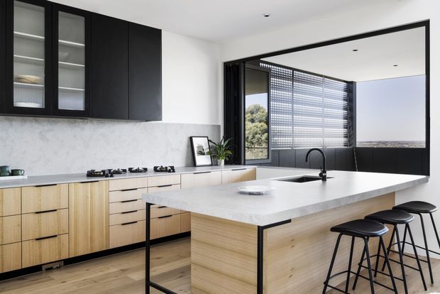 Contemporary kitchen with a large pale stone island, integrated cooktop and sink, light timber cabinetry, and contrasting matte black upper cabinets. Black-framed glazing opens to a bright outdoor view and includes a perforated privacy screen. Three black bar stools line the island, creating a clean, high-contrast, minimalist composition.