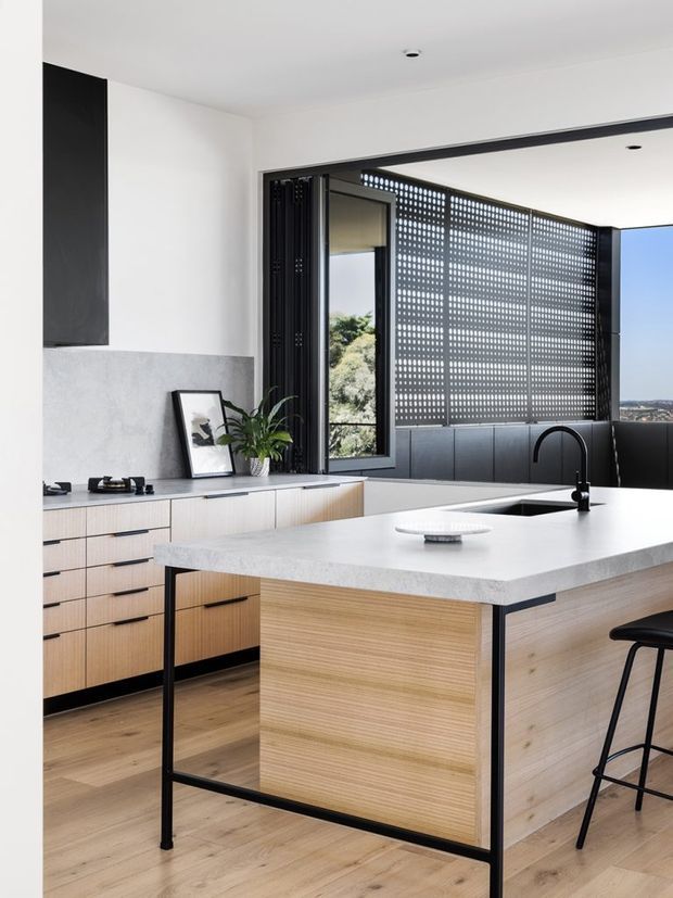 A bright, contemporary kitchen with a large central island, pale timber-grain cabinetry, a light stone benchtop, and slim black metal framing. The room features a black integrated appliance or tall cabinet wall, a black gooseneck tap, a dark tiled or panelled splashback area, and expansive glazing with a perforated privacy screen. Light timber flooring and white walls create an airy, refined interior.