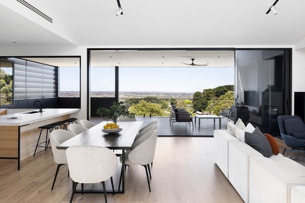 A bright open-plan contemporary interior combining a kitchen, dining area and living room, with pale timber flooring, white cabinetry, a long dining table, upholstered dining chairs and a white sofa. Large black-framed sliding glass openings connect the living space to a covered outdoor terrace and expansive landscaped views. Linear track lighting, minimalist detailing and a ceiling fan reinforce the clean modern design.