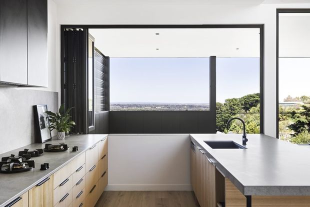 A bright contemporary kitchen with a galley-style arrangement and central circulation space. Light timber lower cabinetry with black pulls, dark upper cabinetry, a pale stone benchtop, integrated gas cooktop, undermount sink, and matte black tap create a clean modern fit-out. Large black-framed windows and an open glazed wall provide expansive views over trees and a distant urban landscape.