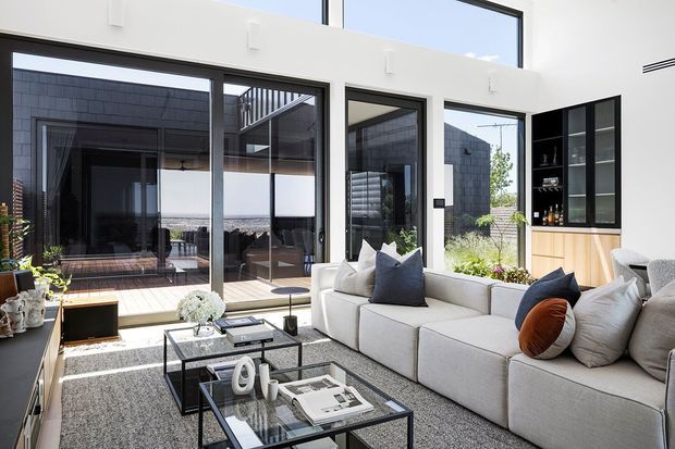 Bright contemporary living room with a large light-coloured sectional sofa, layered cushions, black-framed glass coffee tables, and a textured grey rug. Full-height black-framed glazing opens toward an outdoor courtyard or terrace, while built-in timber-and-black cabinetry with open shelving occupies the right wall. The room has strong indoor-outdoor connection, abundant daylight, and clean-lined detailing.