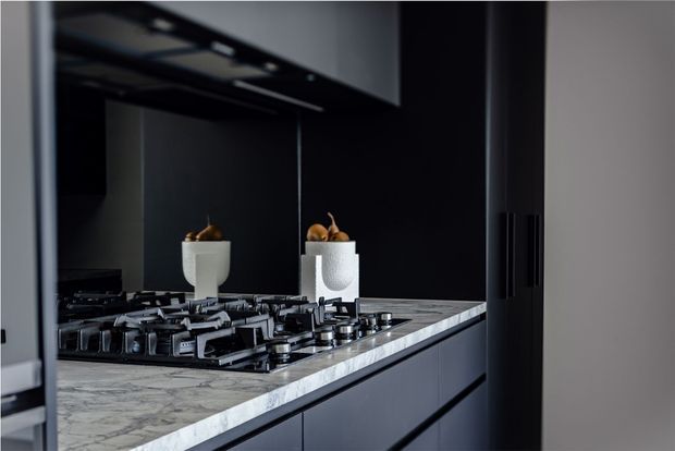 Close-up view of a sophisticated contemporary kitchen, centered on a large gas cooktop set into a veined stone benchtop. Dark matte cabinetry and a dark reflective splashback create a moody, refined look, with minimal decorative objects reflected in the backsplash.