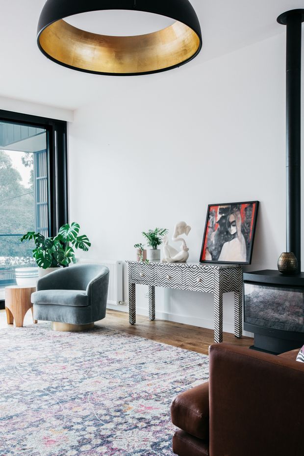 A bright, stylish living room with white walls, timber flooring, a large patterned area rug, and black-framed glazing. A curved grey upholstered armchair sits beside a small timber side table and leafy indoor plants. A striking black-and-white chevron console table supports artwork and sculptural decor, while a black fireplace with a vertical flue occupies the right side. A large black pendant light with a gold interior hangs overhead, and part of a brown leather sofa is visible in the foreground.