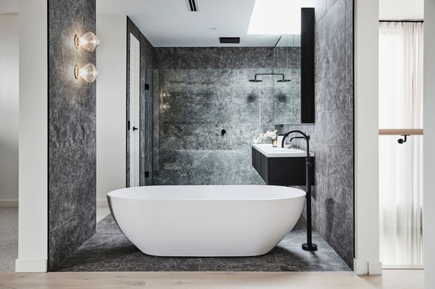 A sophisticated contemporary bathroom with a freestanding white oval bathtub, dark grey stone-look wall and floor finishes, a frameless glass shower enclosure, wall-mounted black fixtures, a floating vanity, and a tall black tap. A bright adjacent area with sheer curtains is visible at the right edge.