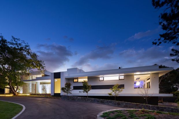 Twilight exterior photograph of a low, elongated modern residence with crisp white horizontal volumes, dark recessed wall sections, narrow clerestory-style windows, and a flat roof. The house sits behind a curved paved driveway with landscaped planting, mature trees, and a low stone retaining edge.
