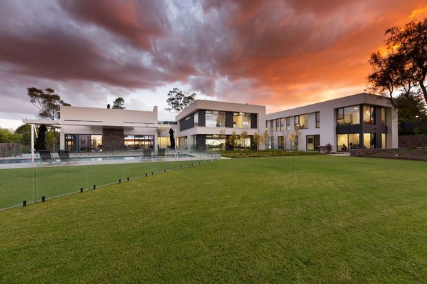 Wide exterior view of a large contemporary multi-wing residence set around a landscaped lawn and swimming pool. The building has clean white and dark grey rendered forms, extensive floor-to-ceiling glazing, flat rooflines, and a transparent pool safety fence. Dramatic sunset clouds dominate the sky, with mature trees surrounding the property.