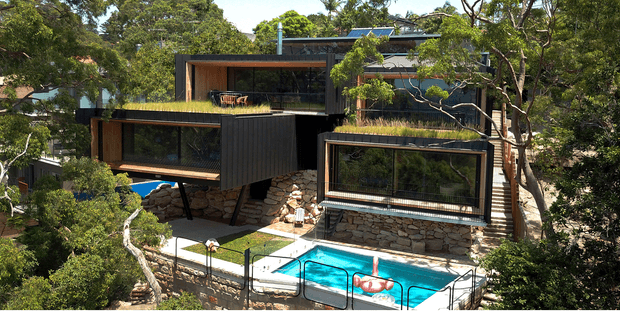 A multi-level contemporary house integrated into a heavily wooded, sloping site. The building features dark cladding, extensive floor-to-ceiling glazing, projecting box-like volumes, planted flat roofs, exposed stone retaining walls, and an outdoor swimming pool in the foreground.