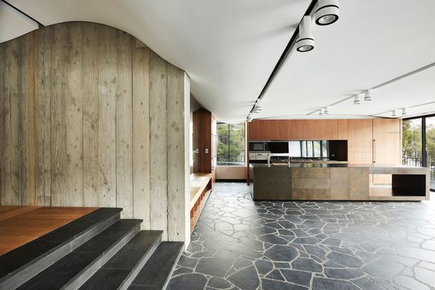 Contemporary open-plan kitchen and living area with a dramatic dark irregular-stone floor, warm vertical timber wall panelling, and a large timber-and-stone kitchen island. The space features sculptural curved ceiling forms, track lighting, extensive glazing, and a short set of dark stair treads leading to a raised timber-floored area.