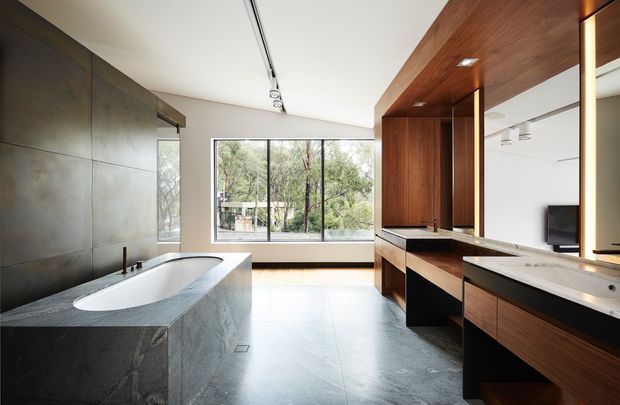 Contemporary bathroom with a freestanding bathtub enclosed in dark stone, extensive timber vanity cabinetry, twin basins, large windows, and integrated track lighting. The space has a clean, spacious, highly refined material palette with views to trees outside.
