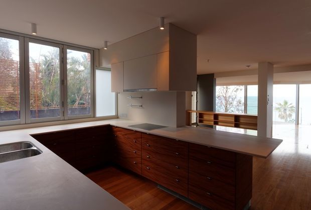 Contemporary open-plan kitchen with a large L-shaped stone-look island and benchtop, dark timber cabinetry, integrated cooktop, stainless sink, and pale overhead cabinetry. Full-height windows and glazing open the space toward a bright coastal outlook, while warm timber flooring and restrained recessed lighting create a refined, airy interior.