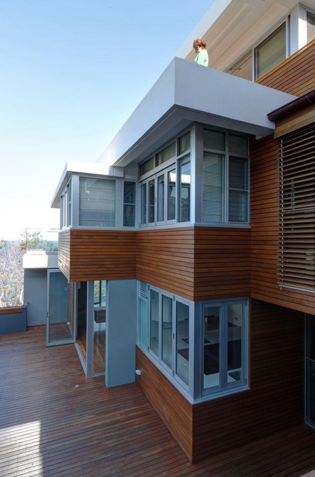Exterior view of a multi-level contemporary residence with projecting box-like volumes, extensive grey-framed glazing, warm horizontal timber cladding, roof terraces, and a broad timber deck. The composition emphasizes the building facade and stepped architectural form, with a glimpse of water and landscape in the distance.