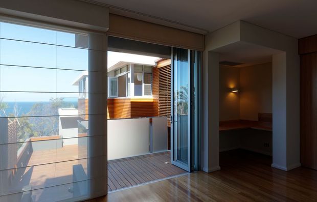 Interior view of a contemporary living area opening onto a timber deck through extensive floor-to-ceiling glazing and sliding glass doors. The room has polished timber flooring, warm neutral walls, a recessed dimly lit built-in nook, and views toward a neighbouring modern timber-clad structure and distant coastal landscape.