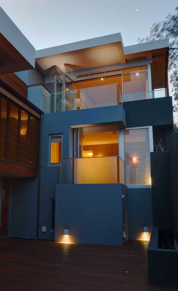 A dusk exterior view of a narrow multi-storey contemporary residence, featuring dark rendered geometric volumes, projecting flat roof planes, large glazed openings, upper-level balconies with clear glass balustrades, warm interior lighting, and a timber deck courtyard. A small crescent moon and surrounding vegetation are visible in the background.