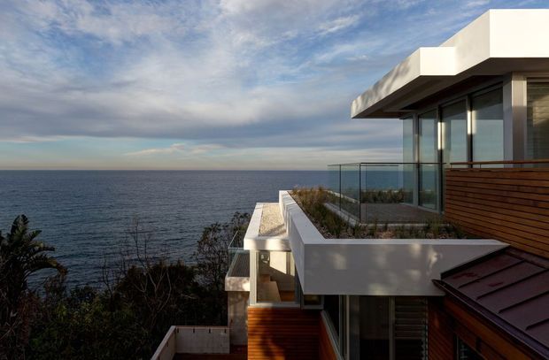 Exterior view of a contemporary multi-level coastal home perched above the ocean, featuring strong horizontal lines, projecting flat roof forms, extensive glazing, glass balustrades, white rendered walls, and warm timber cladding. Terraced roof areas and planted edges step down toward the dramatic coastal setting.