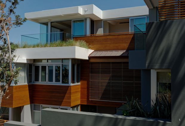 Contemporary multi-level residential facade with crisp white rendered forms, extensive glazing, clear glass balcony balustrades, and warm horizontal timber-look cladding and screening. The building steps around recessed terraces and shaded openings, creating a layered modern exterior composition.
