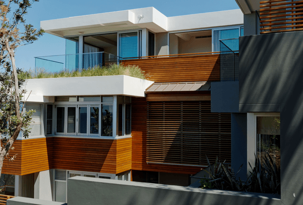 Exterior view of a contemporary multi-level residence with crisp white rendered volumes, extensive glazing, warm horizontal timber cladding and screening, dark metal elements, balconies, and landscaped planting. The composition emphasizes the layered facade and strong geometric forms.