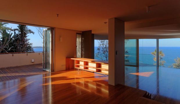 Open-plan modern living area with warm timber flooring, built-in low shelving, broad floor-to-ceiling glazing, and sliding doors opening toward a balcony or terrace. The room overlooks a vivid blue ocean, with strong late-day sunlight creating reflections across the polished floor.
