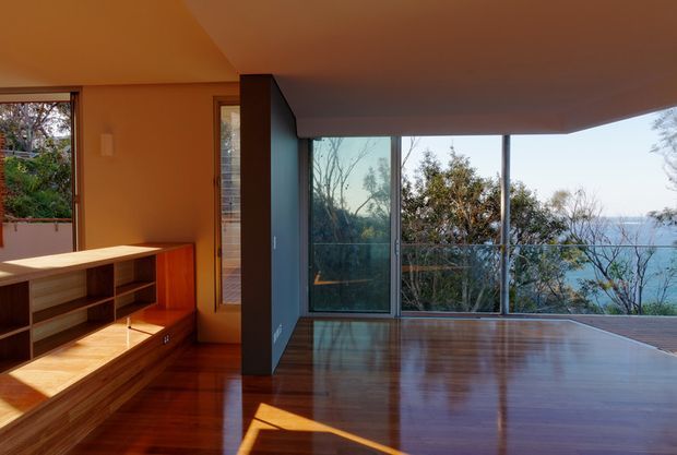 Interior view of a modern living room with extensive floor-to-ceiling glazing overlooking trees and blue water. The room features warm polished timber flooring, a built-in timber media/storage unit, a dark vertical partition, and a low ceiling with a projecting angular overhang. Strong sunlight creates reflections across the floor.