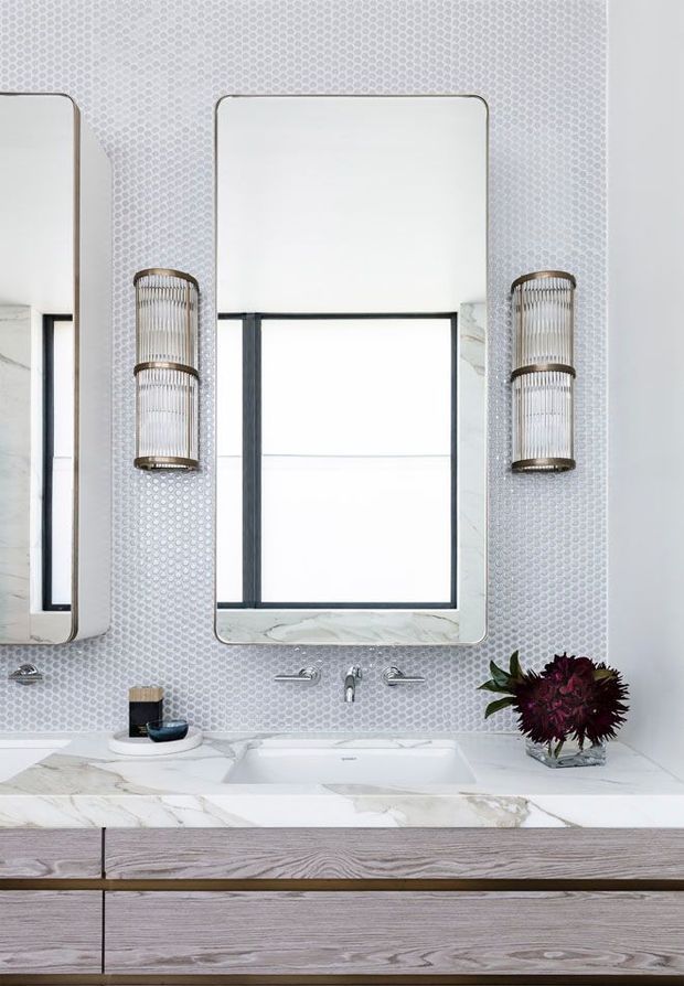 A bright, refined bathroom vanity with a large marble countertop, integrated rectangular basin, wall-mounted chrome tapware, and twin rounded-rectangle mirrors. The wall is finished with small pale textured mosaic tiles, complemented by ribbed metal-and-glass wall sconces and a light timber floating vanity.