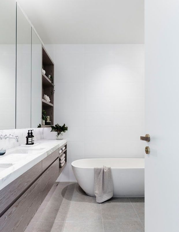 Bright contemporary bathroom viewed through an open door, featuring a freestanding white bathtub, large wall mirror, double vanity with pale wood-grain cabinetry and white countertop, open shelving, white tiled walls, and large grey floor tiles. A folded towel and simple black bathroom accessories add subtle contrast.