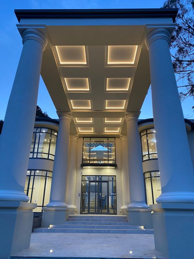 Formal grand entrance to a large classical-style building, viewed from the front at dusk. A deep portico is supported by tall round columns with ornate moulded capitals and bases, beneath a coffered ceiling with recessed illuminated panels. Broad steps lead to glazed double entry doors, with curved multi-storey wings and dark-framed windows on either side.