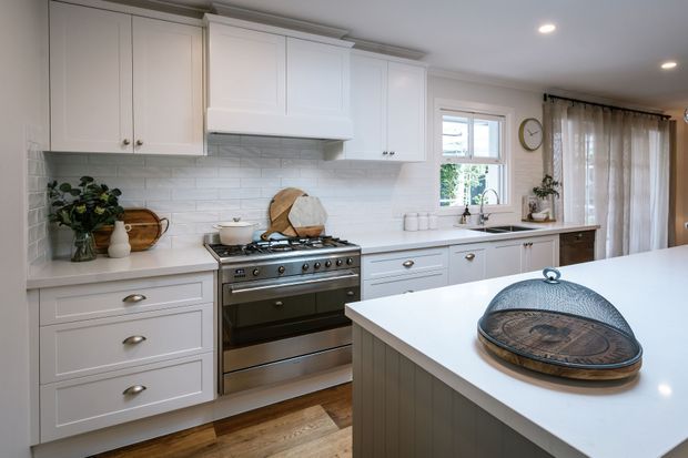 Bright, spacious traditional-style kitchen with white shaker cabinetry, a large central island, stainless-steel range cooker, white subway-tile splashback, and warm timber flooring. Natural light enters through the window and glazed door, with neutral furnishings and brushed metal hardware creating a refined, welcoming look.