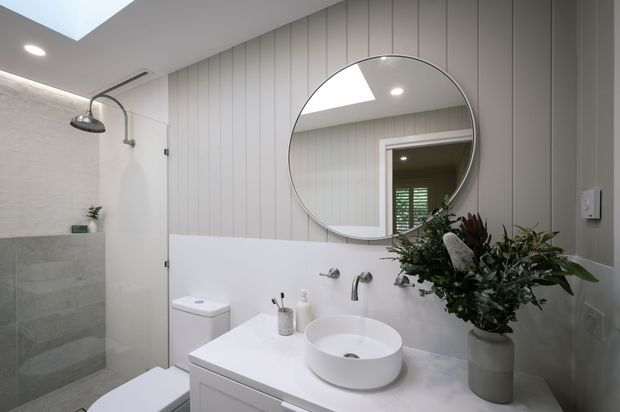 A bright, contemporary bathroom featuring a white floating vanity with a round vessel basin, wall-mounted tapware, a large circular mirror, vertical grey wall panelling, and a walk-in shower with pale grey tiled walls. A skylight and recessed lighting provide natural and ambient illumination, while greenery adds a soft organic accent.